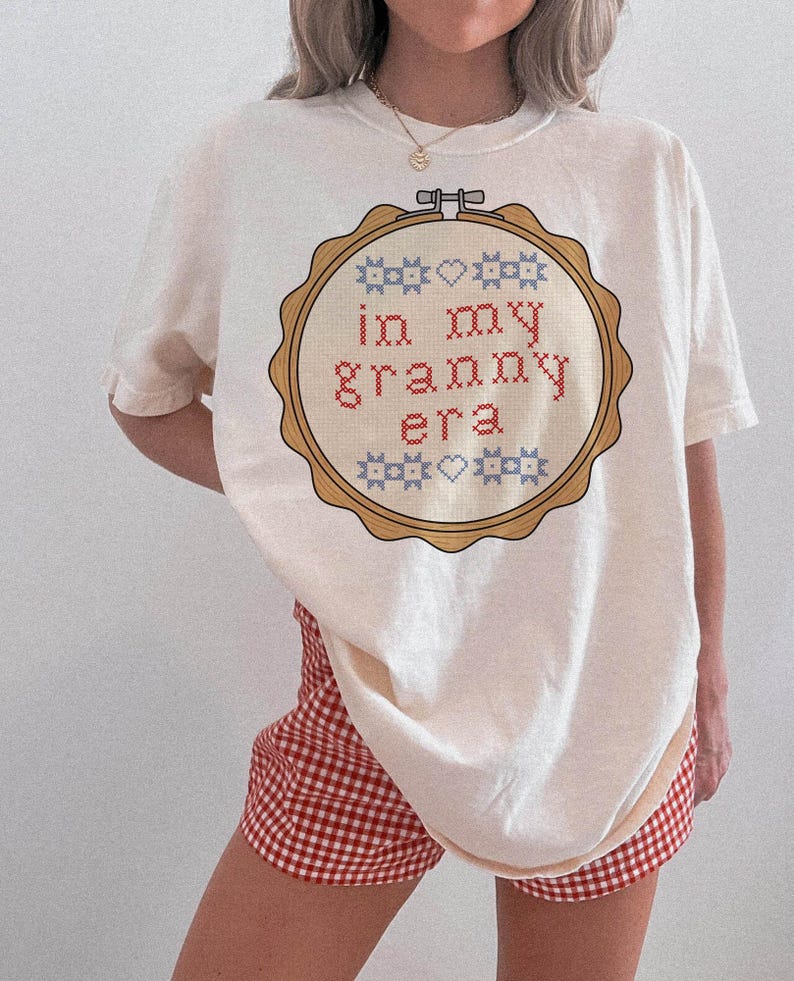 Puede incluir: Camiseta color crema con un dise&ntilde;o de punto de cruz dentro de un bastidor de madera. El dise&ntilde;o incluye el texto "in my granny era" en rojo, con elementos decorativos azules y blancos. La modelo lleva un pantal&oacute;n corto a cuadros rojos y blancos.