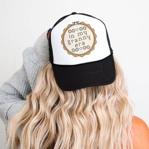 Puede incluir: Gorra de camionero en blanco y negro con el texto "in my granny era" dentro de un dise&ntilde;o de aro de bordado beige. La gorra tiene una visera negra y un panel frontal blanco. La persona lleva el pelo largo y rubio.