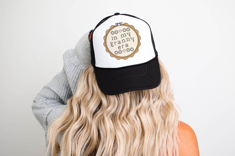 Puede incluir: Gorra de camionero en blanco y negro con el texto "in my granny era" dentro de un dise&ntilde;o de aro de bordado beige. La gorra tiene una visera negra y un panel frontal blanco. La persona lleva el pelo largo y rubio.