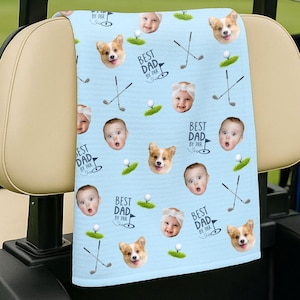 May include: A light blue golf towel featuring a repeating pattern of baby faces, corgi faces, golf clubs, and golf balls. The text "BEST DAD BY PAR" is printed on the towel. The towel is draped over the back of a golf cart.