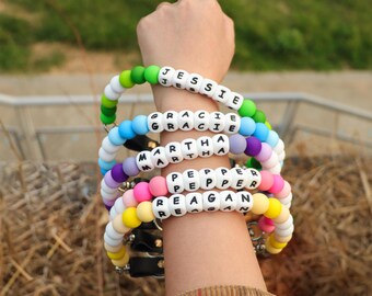 May include: A stack of colorful beaded bracelets spelling out names: Jessie, Gracie, Martha, Pepper, and Reagan. The bracelets feature white letter beads and a variety of colored beads, including green, blue, purple, pink, and yellow.