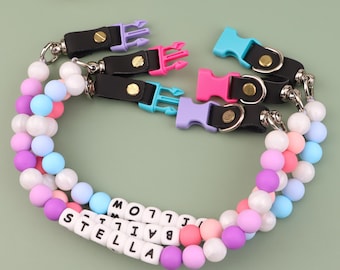 May include: A beaded pet collar with the name "STELLA" spelled out in white letter beads. The collar features a variety of pastel-colored beads and black leather straps with colorful plastic buckles in purple, pink, and blue.