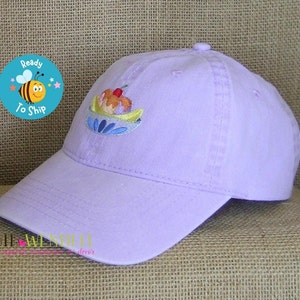 May include: A light purple baseball cap with a curved brim. The cap features an embroidered design of a sundae with a cherry on top. A circular sticker with a bee and the words "Ready To Ship" is visible.