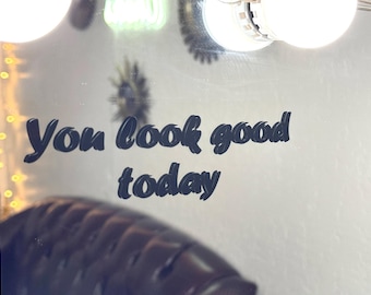 May include: A rectangular mirror featuring the black text "You look good today" in a bold, stylized font. The mirror surface reflects a dark tufted piece of furniture and is illuminated by bright, round light bulbs positioned along the top edge.