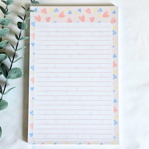 Pode incluir: Um bloco de notas com linhas com uma borda de padrão de coração pastel. Os corações são em rosa, azul e amarelo. O bloco de notas tem um fundo rosa claro e linhas horizontais vermelhas para escrever. O bloco de notas é retangular.