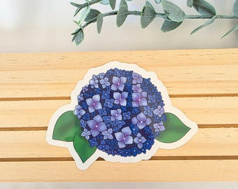 Puede incluir: Una pegatina con una ilustración detallada de una flor de hortensia azul y morada con hojas verdes. La pegatina está sobre una superficie de madera con una ramita de vegetación en el fondo. El diseño floral es vibrante y colorido.