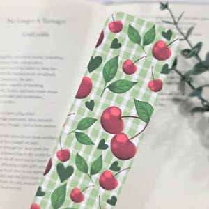 May include: A decorative bookmark featuring a green and white gingham pattern. The bookmark is adorned with illustrations of red cherries, green leaves, and green hearts. The bookmark is placed on top of an open book.