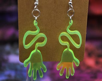 May include: A pair of green acrylic earrings shaped like hands with a wavy line extending from the wrist. The earrings are on a brown card.