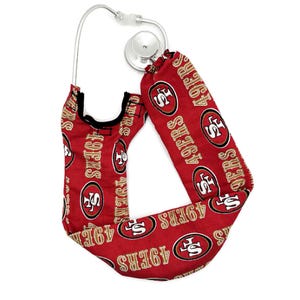 May include: A stethoscope cover made of red fabric featuring a repeating pattern of the San Francisco 49ers logo and the text 49ERS in gold and white. The cover is designed to wrap around the tubing of a standard medical stethoscope.