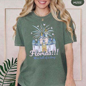 K&ouml;nnte beinhalten: Moosgr&uuml;nes T-Shirt mit einer Illustration eines M&auml;rchenschlosses, Feuerwerk und dem Text "Florida!!! one hell of a drug." Das Design beinhaltet Mickey Mouse Silhouetten. Dieses kurz&auml;rmlige T-Shirt ist ein Freizeitkleidungsst&uuml;ck.
