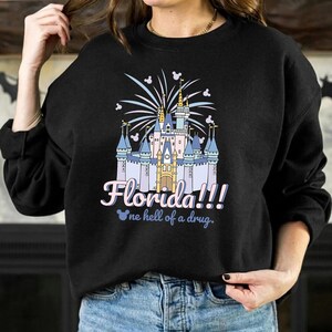 K&ouml;nnte beinhalten: Schwarzes Sweatshirt mit einer farbenfrohen Grafik eines Schlosses, Feuerwerk und dem Text "Florida!!!" in Rosa und Blau. Darunter steht der Satz "one hell of a drug" in Hellblau.