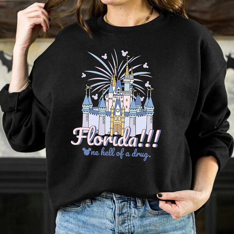 K&ouml;nnte beinhalten: Schwarzes Sweatshirt mit einer farbenfrohen Grafik eines Schlosses, Feuerwerk und dem Text "Florida!!!" in Rosa und Blau. Darunter steht der Satz "one hell of a drug" in Hellblau.