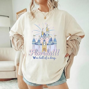 K&ouml;nnte beinhalten: Cremefarbenes T-Shirt mit einer farbenfrohen Illustration eines Schlosses und dem Text "Florida!!!" in Schreibschrift. Darunter der Satz "one hell of a drug". Das Schlossdesign hat blaue, rosa und goldene Akzente.