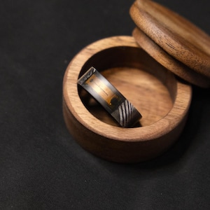 Könnte beinhalten: Ein handgefertigter Ring mit einzigartigem Design liegt in einer kleinen Holzschale. Der Ring hat ein dunkles Band mit einem Mittelteil in Gold- und Bernsteintönen und einen gemusterten silber-schwarzen Abschnitt.