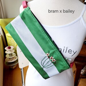 Op de afbeelding: Een groene en witte gestreepte stoffen tas met een roze band en geborduurde bloemen. De tas heeft een ritssluiting en de tekst "bram x bailey" is op de stof gedrukt.
