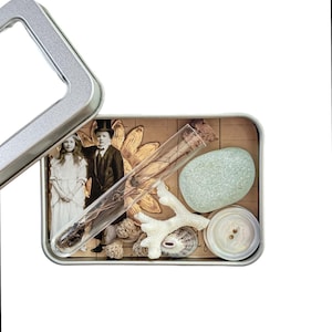 Könnte beinhalten: Eine Vintage-Blechdose gefüllt mit verschiedenen Gegenständen, darunter ein Reagenzglas aus Glas, eine Muschel, ein weißer Knopf, ein kleines Stück Koralle, ein glatter hellblauer Stein und ein Vintage-Foto eines Paares.