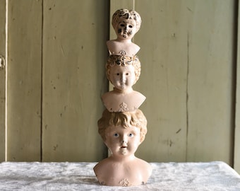May include: Three antique bisque doll heads stacked on top of each other. The heads are painted in a pale pink color with detailed facial features and curly blonde hair. The heads are displayed on a white cloth.