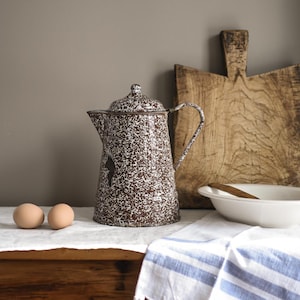 Puede incluir: Una cafetera de esmalte moteado marrón y blanco con tapa y asa. Dos huevos marrones están sobre un paño blanco frente a la cafetera. Una tabla de cortar de madera y un cuenco blanco están en el fondo.