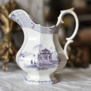 May include: White ceramic pitcher with a detailed purple landscape design. The pitcher has a curved handle and a decorative rim. The landscape scene features buildings, figures, and trees. A classic piece for serving beverages.