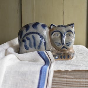 Puede incluir: Figura de gato de cerámica gris y azul, sobre un paño blanco con rayas azules. El gato tiene un diseño estilizado con marcas azules y una cara detallada. La figura es un objeto decorativo.