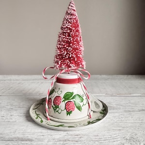 May include: A decorative Christmas tree sits atop a vintage teacup and saucer set. The red tree is adorned with white accents and tied with a red and white string. The teacup and saucer are white with a strawberry design and green leaf accents.