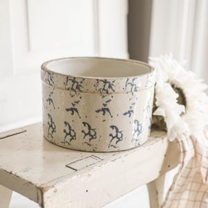 Pode incluir: Um pote de cerâmica cilíndrico com uma base de cor bege claro e um padrão repetitivo de motivos abstratos de flores ou folhas em azul escuro. O recipiente tem um acabamento liso e vidrado com uma borda espessa e resistente.