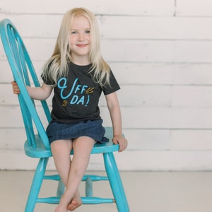 May include: A young girl wearing a dark grey t-shirt with the text "Uff Da!" in blue and yellow lettering. She is sitting on a blue chair against a white wood panel background.