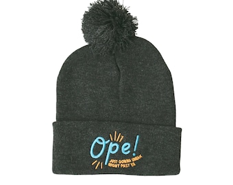 Puede incluir: Un gorro de lana gris oscuro con un pompón negro y el texto "Ope! Just gonna sneak right past ya" bordado en azul y naranja.