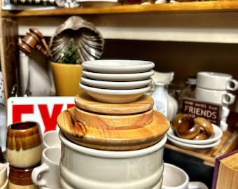 May include: A cylindrical off-white ceramic canister with horizontal ribbed detailing, topped with a stack of three small matching ceramic saucers resting on a circular wooden lid. The canister has a smooth, glossy finish and a neutral color.