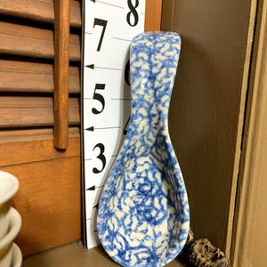May include: A blue and white ceramic spoon rest with a speckled pattern. The spoon rest is teardrop-shaped with a curved handle. The background includes a wooden shutter and a ruler with black numbers.