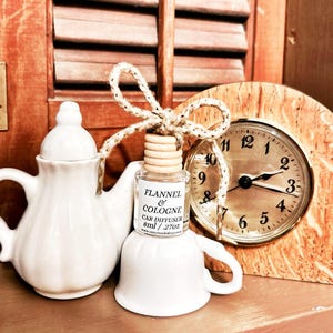 May include: A white ceramic teapot and teacup-shaped car air freshener with a bottle of "Flannel & Cologne" fragrance. A small wooden clock with a gold-colored face and black hands is also present. The clock shows the time as 2:00.