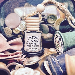 May include: A clear glass car air freshener with a wooden cap and a rope string, labeled "Fresh Linen." The image includes various sewing notions like buttons, spools of thread, and fabric scraps in a vintage-style setting.