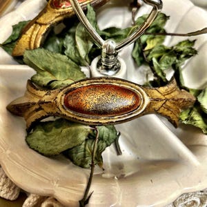 May include: A decorative drawer pull featuring an elongated, ornate gold-toned metal frame with a distressed finish. The center of the pull contains an oval-shaped inlay with a gradient of deep red to orange, speckled with dark brown flecks.