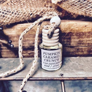 May include: A small glass car air freshener with a wooden top and a rope for hanging. The label reads "Pumpkin Caramel Crunch Car Diffuser". The diffuser is 8ml / .27oz.