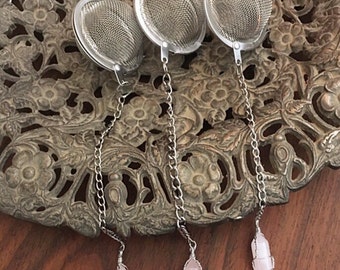 May include: Three silver tea infusers with chains and pink crystal charms. The infusers are round with a fine mesh and are hanging from a silver chain. The charms are pink quartz crystals.