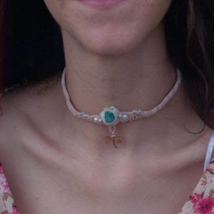 Può includere: Una collana girocollo in cordino intrecciato rosa chiaro con una pietra ovale verde centrale, piccole perle bianche e perline azzurre. Un piccolo ciondolo a forma di libellula metallica pende dal centro del cordino intrecciato sul collo.