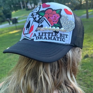 May include: Black and white trucker hat with a mesh back. The front panel is white and decorated with playing cards, hearts, and a sequined rose. The text "A Little Bit Dramatic" is displayed on the brim.