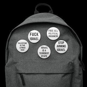 Könnte beinhalten: Ein grauer Rucksack mit mehreren weißen Ansteckern. Die Anstecker tragen schwarzen Text, darunter "FUCK ISRAEL", "PALESTINE IS THE LITMUS TEST", "FREE ALL POLITICAL PRISONERS", "ISRAEL IS A TERRORIST STATE" und "STOP ARMING ISRAEL".