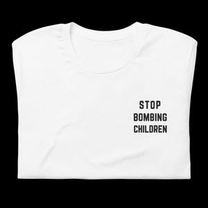 Op de afbeelding: Gevouwen wit T-shirt met de zwarte tekst "STOP BOMBING CHILDREN". De tekst staat rechts op het shirt. Het T-shirt wordt gepresenteerd tegen een zwarte achtergrond.