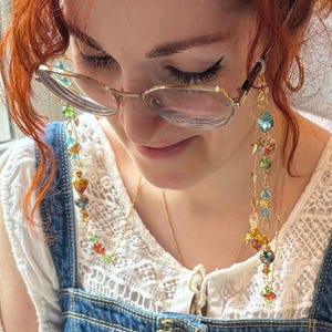 May include: A gold-toned eyeglass chain featuring a variety of colorful beads, including translucent blue, green, and orange spheres, along with small gold-toned leaf charms and floral-patterned beads, attached to gold-toned metal eyeglass frames.