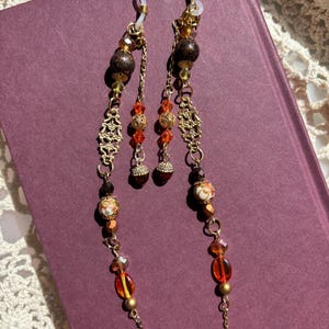 May include: A pair of eyeglass chains featuring gold-tone metal links, clear silicone loops, and a variety of beads including dark brown spheres, floral patterned ceramic beads, faceted orange crystals, and small metallic acorn-shaped charms.