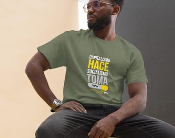 May include: Olive green t-shirt with the text "CAPITALISMO HACE SOCIALISMO TOMA" in white and yellow. The shirt is worn by a person sitting on a black stool, wearing black jeans and white sneakers.