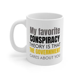 Könnte beinhalten: Weiße Keramik-Kaffeetasse mit gebogenem Henkel. Die Tasse zeigt den Text "My favorite CONSPIRACY THEORY IS THAT THE GOVERNMENT CARES ABOUT YOU" in schwarzer, grauer und gelber Schrift.