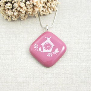 May include: A pink, square pendant necklace with a silver chain. The pendant features a white birdhouse design with a clock face, birds, and branches. The necklace is a delicate piece of jewelry.