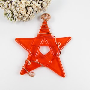 May include: Orange glass star ornament with copper wire wrapped around it. The star has a hole in the center and is a festive decoration.