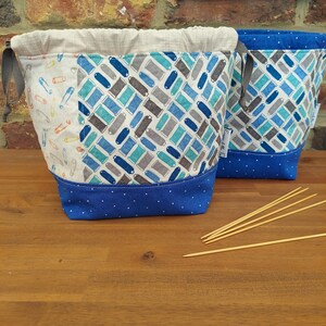 Könnte beinhalten: Zwei Stoff-Projektbeutel mit Kordelzugverschlüssen. Eine Tasche hat ein Muster aus blauen, grauen und türkisfarbenen Spulen, während die andere ein Muster aus Sicherheitsnadeln hat. Beide Taschen haben einen blauen Tupfenboden und ein neutrales Futter.