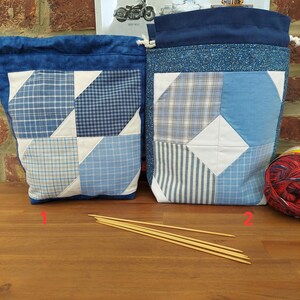 Op de afbeelding: Twee blauwe patchwork-taszakken met trekkoord en witte accenten. De tassen hebben verschillende blauwe ruitjes- en patroonstoffen. Houten breinaalden staan op de voorgrond. Een bol wol is rechts zichtbaar.