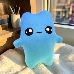 Peut inclure: Une peluche bleu clair avec une texture douce et lisse. Le personnage a une forme de corps simple et arrondie avec deux petits bras et deux jambes. Il présente de grands yeux noirs circulaires avec des reflets blancs et une petite bouche noire.