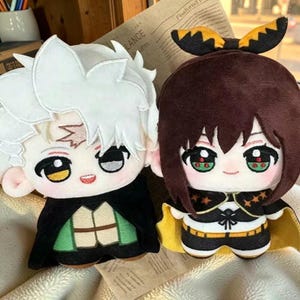 Könnte beinhalten: Zwei Plüschpuppen im Anime-Stil. Die linke Puppe hat weißes Haar, einen schwarzen Umhang und eine grüne Weste. Die rechte Puppe hat braunes Haar mit einer schwarz-gelben Schleife, einen schwarzen Umhang mit gelbem Futter sowie ein schwarz-weißes Outfit.