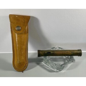 May include: A cylindrical brass pocket level tool with a visible bubble vial and engraved text, accompanied by a tan leather protective sheath featuring a metal snap closure. The tool rests horizontally across a clear glass ashtray.